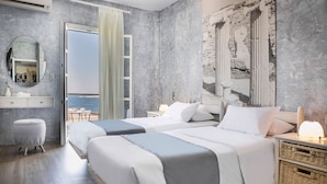 Quarto de hotel com duas camas, um espelho grande, um banquinho pequeno, uma mesa de cabeceira com abajur e vista para o mar através de uma porta aberta.