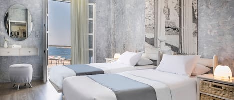 Hotellihuone, jossa on kaksi sänkyä, suuri peili, pieni jakkara, yöpöytä lampun kanssa ja näkymä merelle avoimesta ovesta.