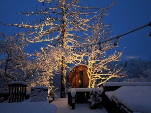 Un espacio al aire libre cubierto de nieve, con una cabaña de madera, bancos y árboles cargados de nieve.