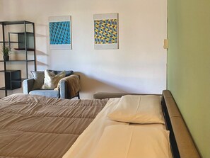 A hotel room with a bed, a chair, a small table, and two framed artworks on the wall.