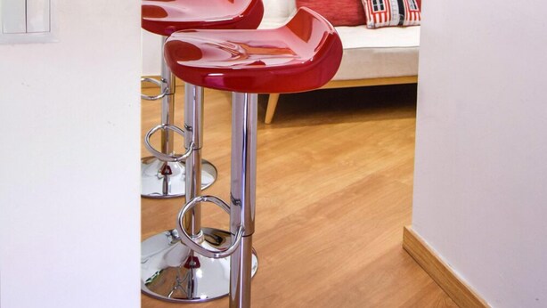 Una habitación moderna con suelo de madera, una pared blanca con obras de arte, un sofá rojo y blanco, y un taburete alto rojo.
