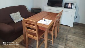 Een woonkamer met een bruine bank, een houten eettafel met vier stoelen, een flatscreen televisie, een magnetron en een klein kastje.