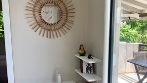 Rincón de una habitación con pared blanca, un adorno de pared de bambú decorativo, un jarrón blanco con ramas y un estante pequeño con libros y un búho decorativo.