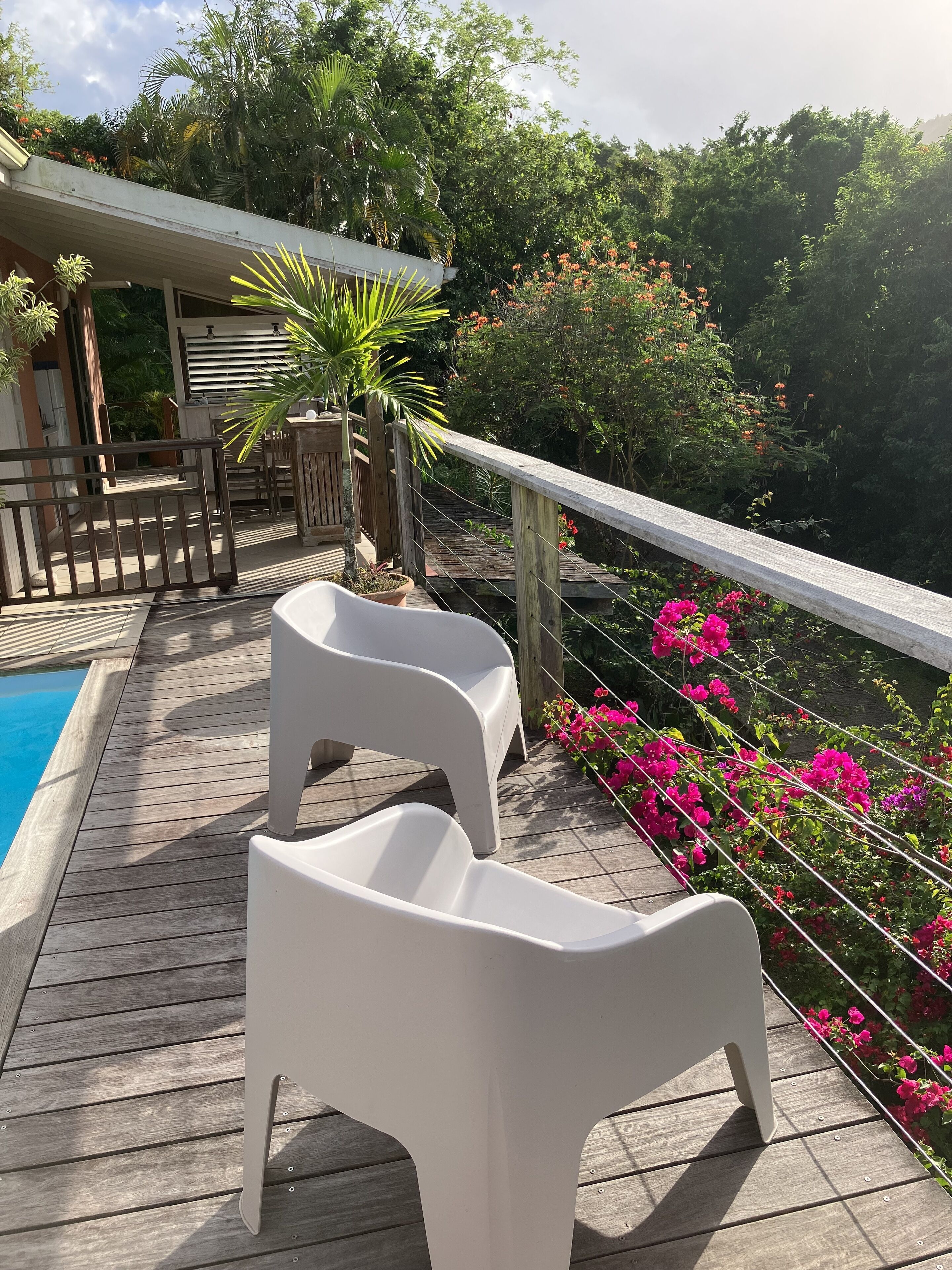 A wooden deck with two white chairs, a pool, and a view of lush greenery and flowers.