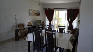 Un coin repas avec une table dressée pour quatre personnes, entourée de chaises dont les dossiers sont en osier tressé. Un téléviseur est fixé au mur, et un vase contenant des plantes séchées se trouve sur le sol.