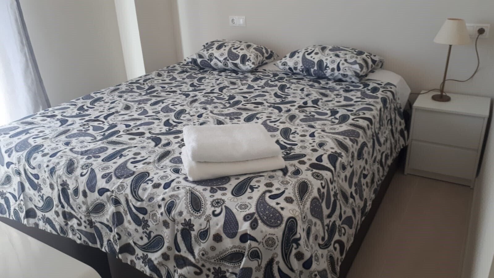 A neatly made bed with a patterned bedspread and pillows, a white bedside table with a lamp, and a plain wall.
