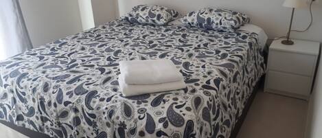 A neatly made bed with a patterned bedspread and pillows, a white bedside table with a lamp, and a plain wall.