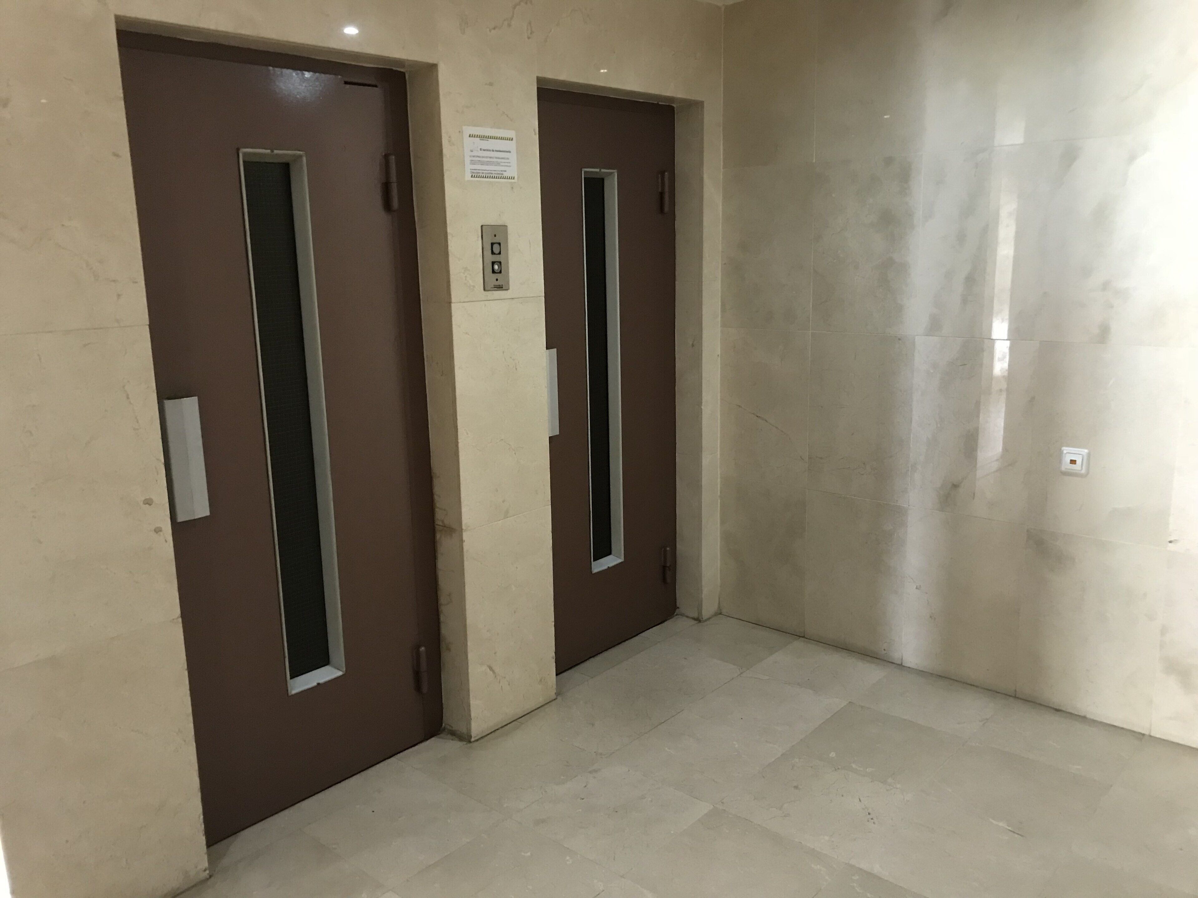 A hallway with two elevator doors, a light switch, and a wall-mounted sign.