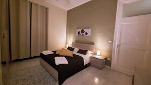 A neatly made bed with pillows and a bedside table with a lamp, a wall with a floral artwork, and a closed door. - LA MAISON SR Cozy apartment in the center of town (Campobasso)