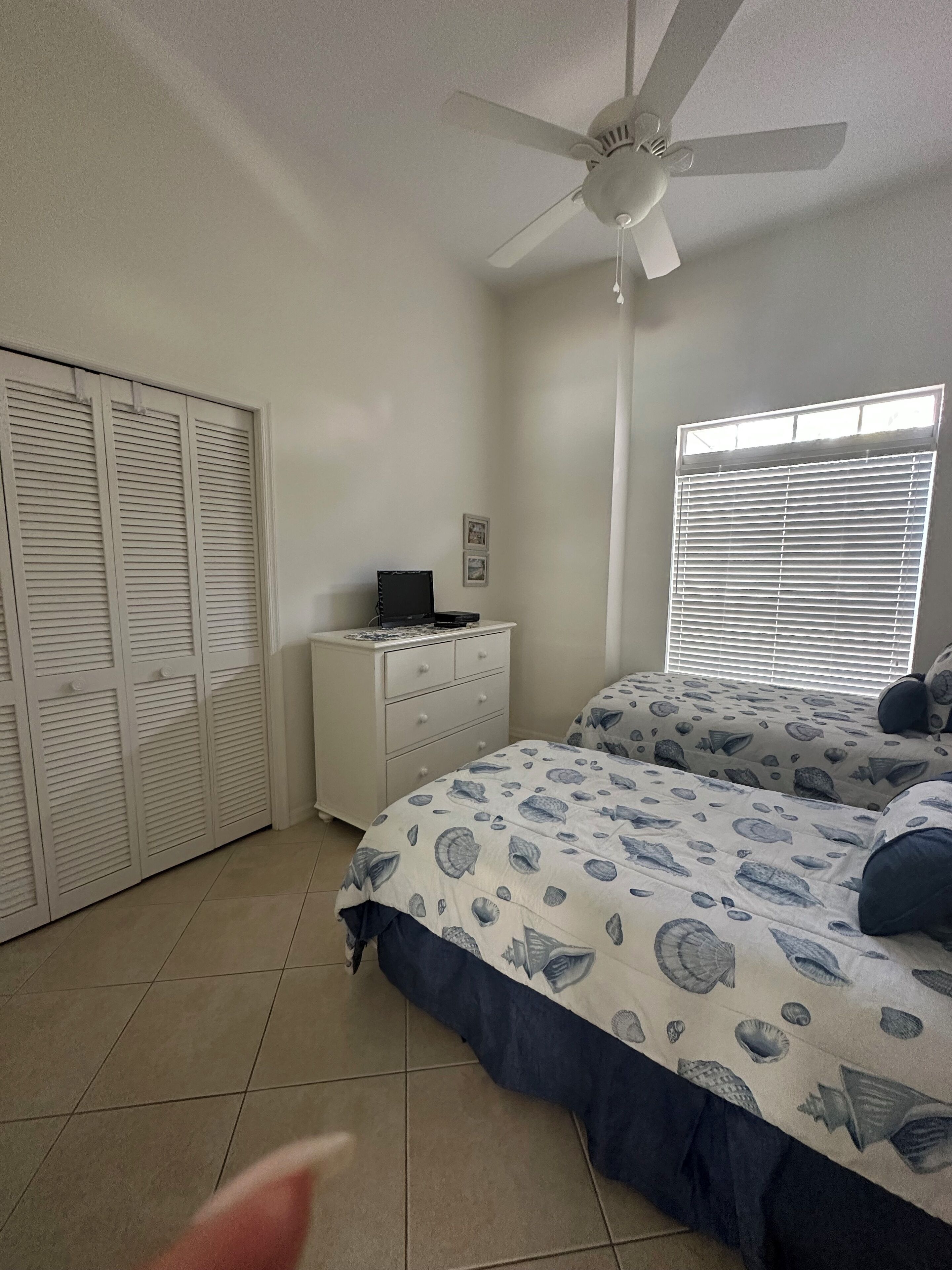 A bedroom with a ceiling fan, a window with blinds, a bed with a patterned bedspread, and a dresser with a laptop.