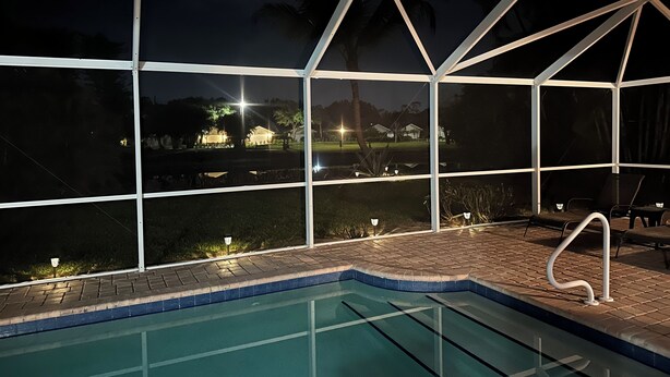 Una piscina con cerramiento de vidrio, un área junto a la piscina con sillas y vistas a un parque con árboles y un edificio al fondo.