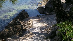 Una costa rocosa con aguas transparentes, un sendero arenoso y abundante vegetación.