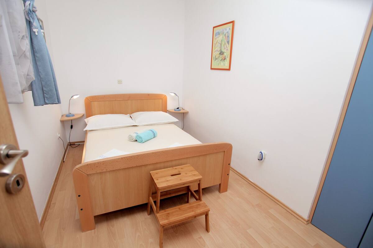 A bedroom with a wooden bed, bedside tables, a small wooden stool, a wall-mounted picture, and a door with a handle.