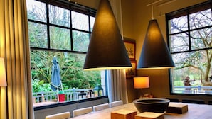 A modern dining area with a long wooden table, two black pendant lamps, and a view of the outdoors through large windows. - House to rent for 6-12 people in Vaudreuil-27-Louviers-Gaillon- Andé-Rouen (connelles)