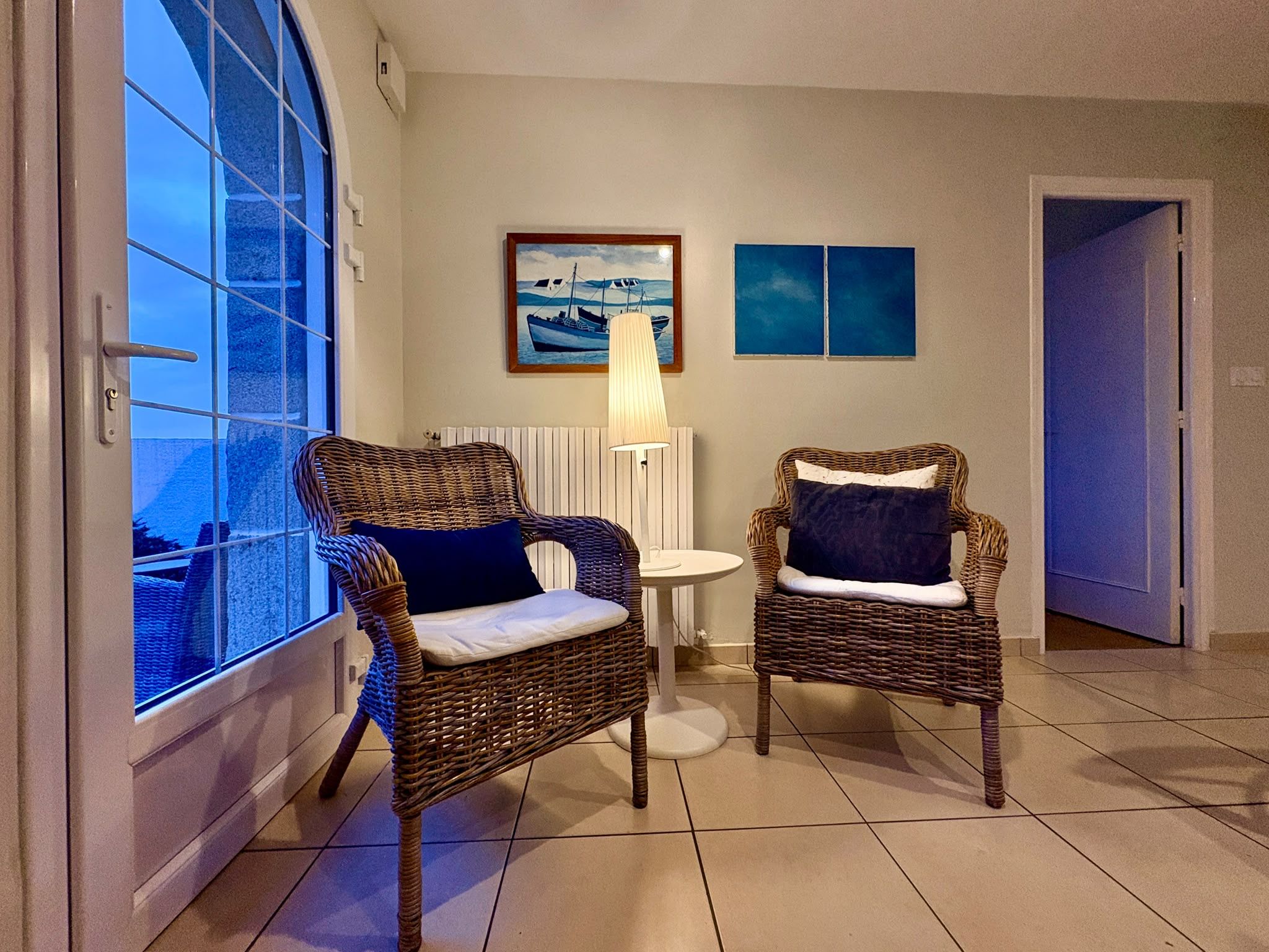 A room with two wicker chairs, a table, a lamp, and a painting on the wall.