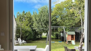 Puerta corrediza de vidrio que da a un patio exterior con terraza de madera, sillas de plástico blanco, una casita de juegos y un columpio.