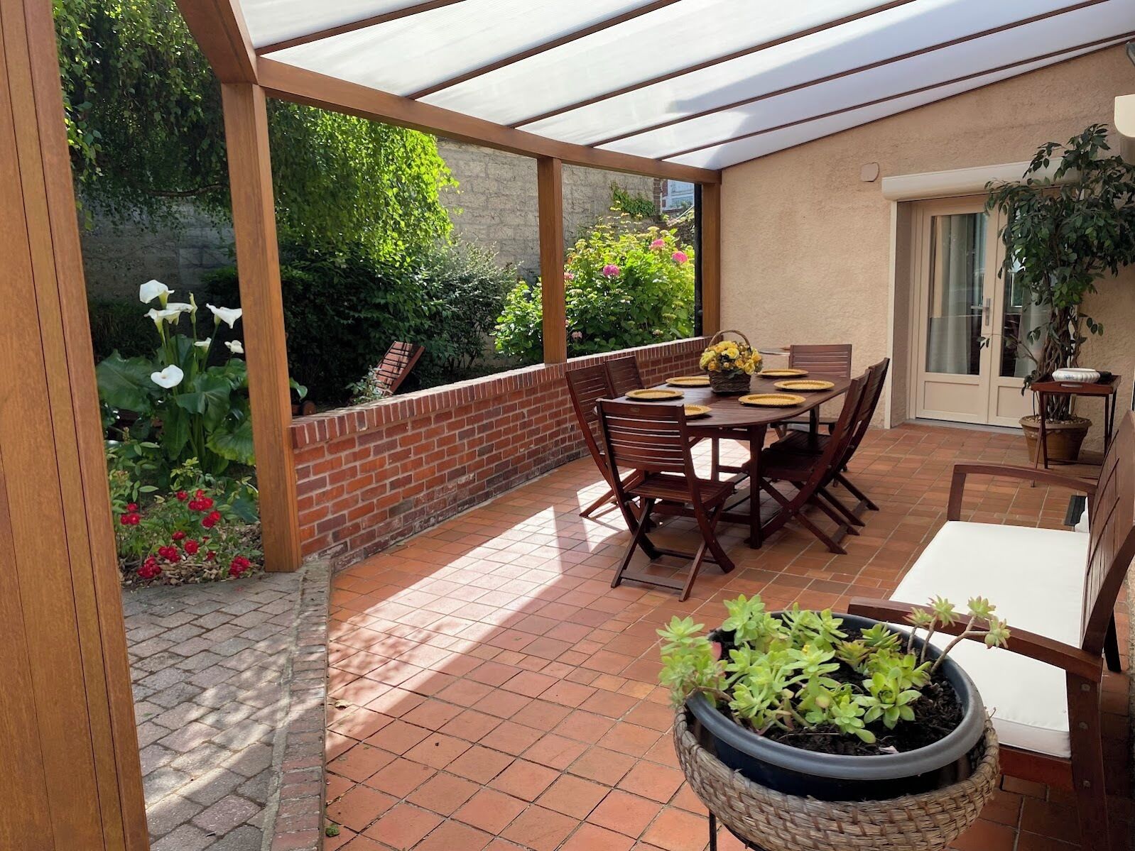 A covered patio with a brick floor, a wooden table with chairs, and a potted plant.