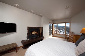 A bedroom with a fireplace, a large bed, a TV, and a view of a snowy landscape through a large window.