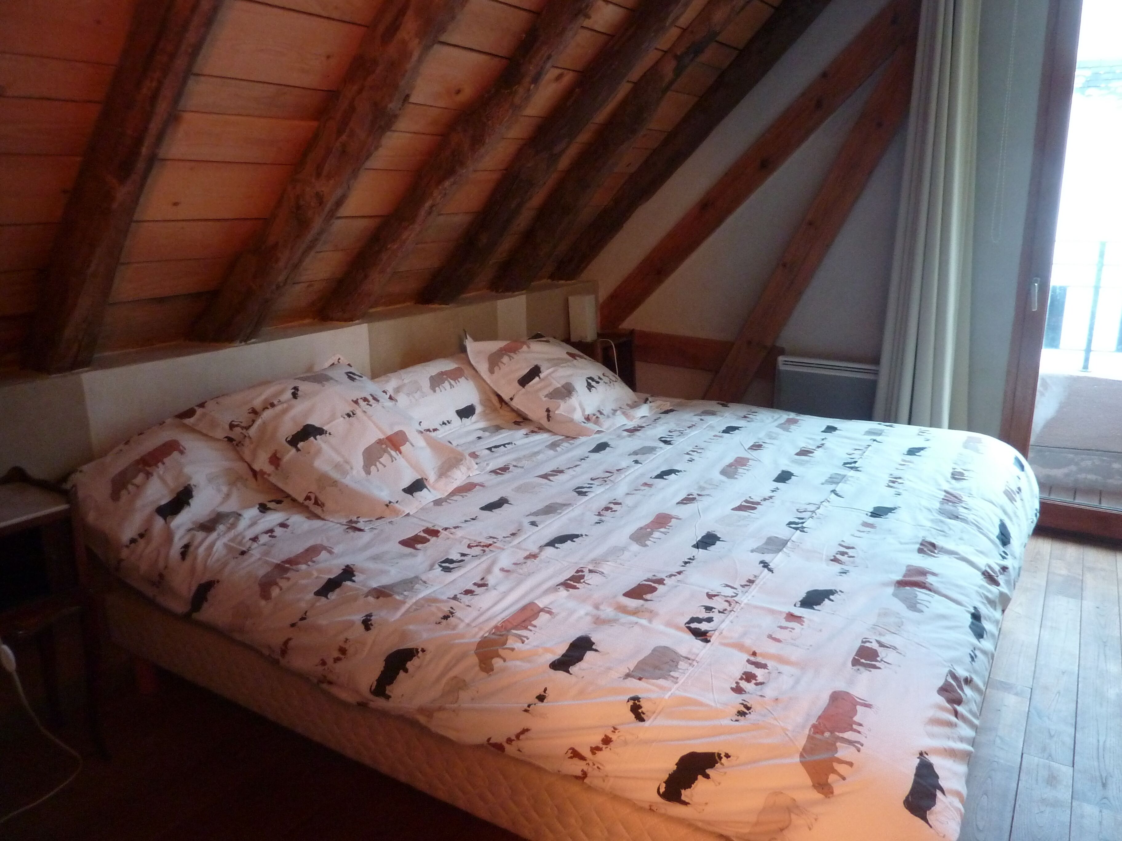 A bedroom with a sloped ceiling, wooden beams, a bed with patterned bedding, and a window with curtains.