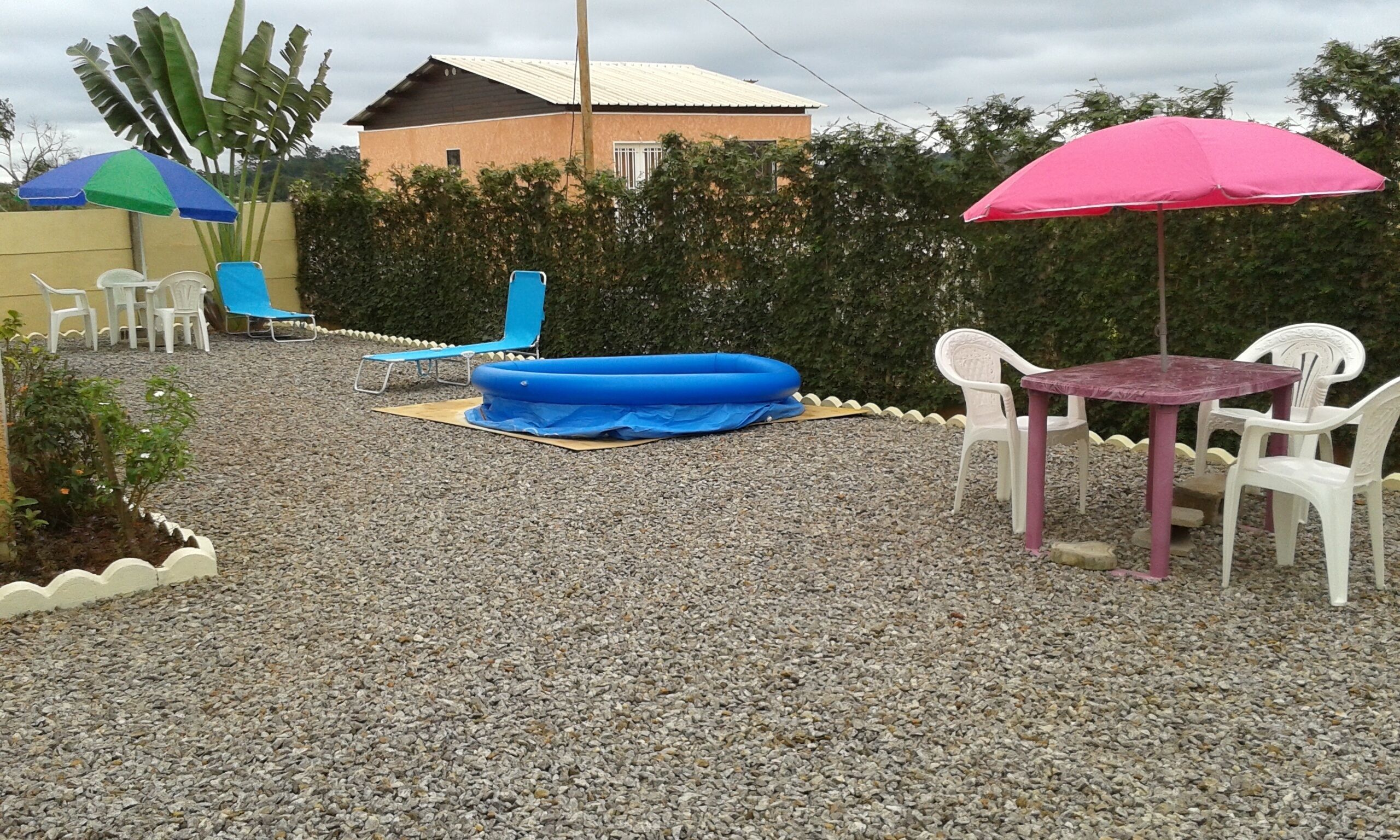 Un espace extérieur aménagé en terrasse, avec une piscine gonflable bleue, des chaises en plastique blanches, une table avec un parasol rose et un bâtiment en arrière-plan.