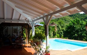 An outdoor pool area with a covered patio, surrounded by lush greenery and a mountainous backdrop.