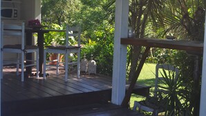 Une terrasse en bois, agrémentée de chaises blanches et d’une table, entourée d’une végétation luxuriante et offrant une vue sur un jardin.