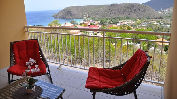 Un balcon avec deux fauteuils en osier et une table, d’où l’on peut admirer une ville côtière avec une montagne en arrière-plan.