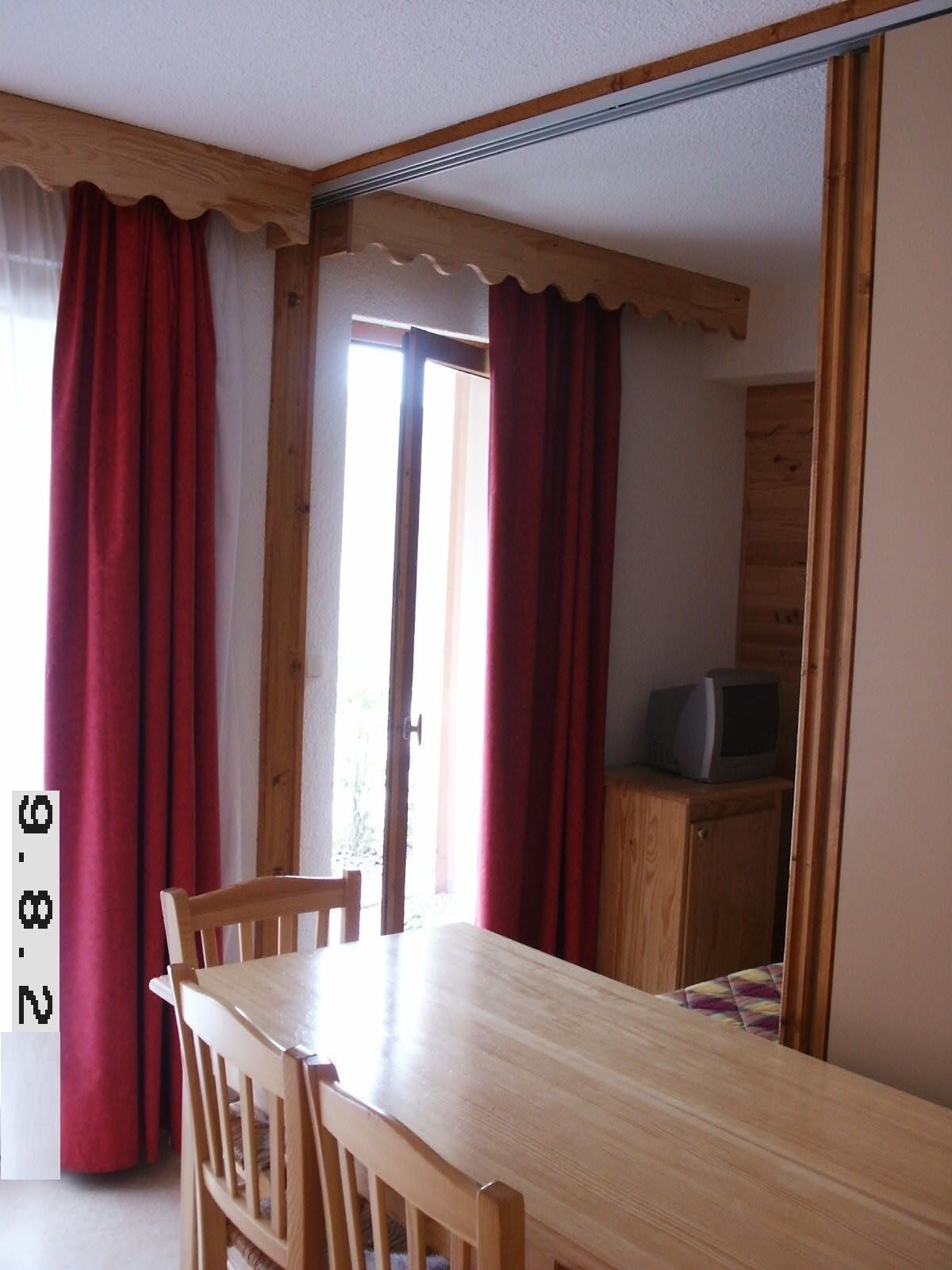 A room with a wooden dining table and chairs, a television on a wooden cabinet, and red curtains.
