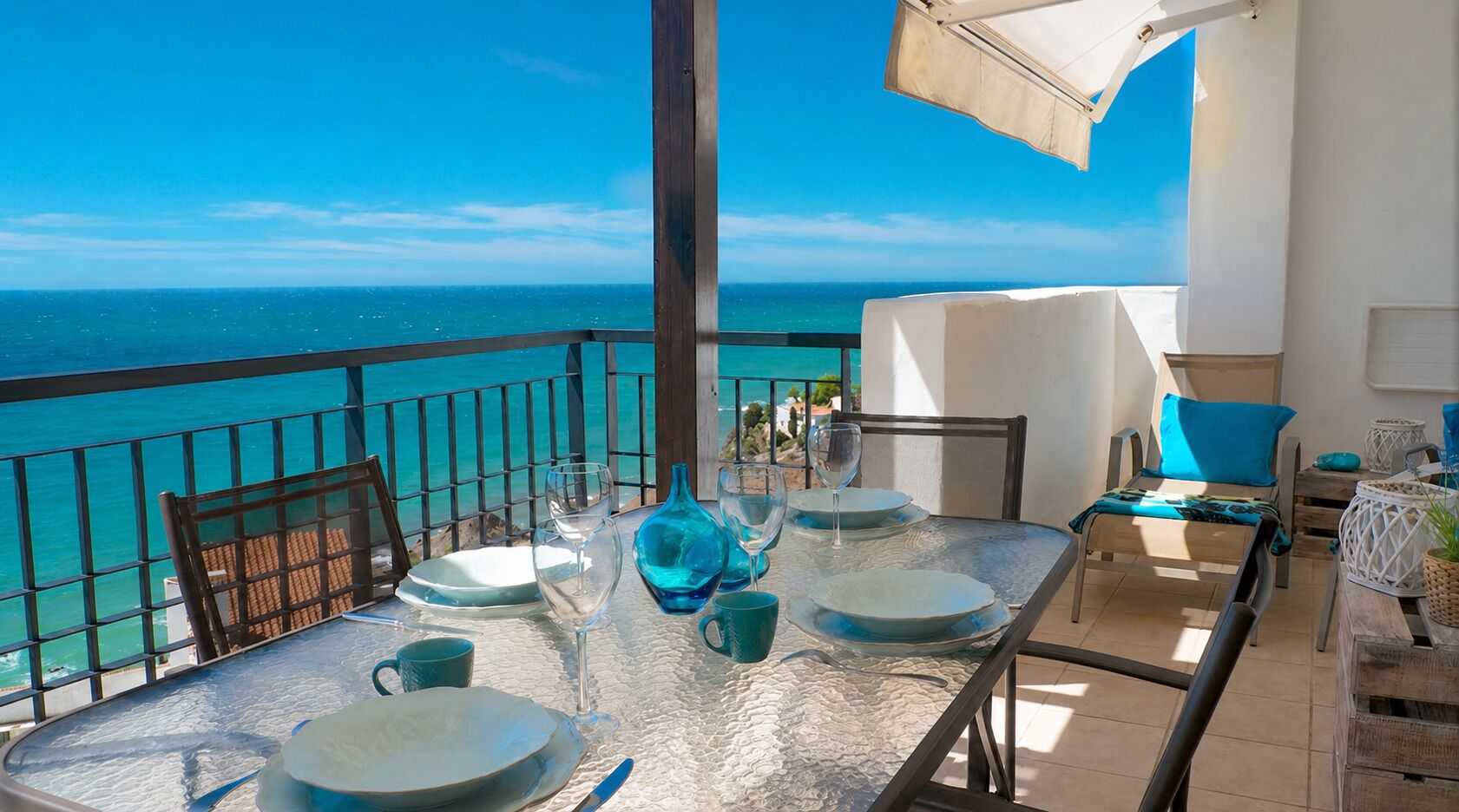 Un balcón con una mesa lista para comer, con vistas al mar azul y despejado.