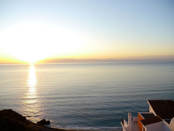 Blick auf die Küste mit Sonnenuntergang über dem Meer, eine felsige Küstenlinie und Gebäude mit weißen Wänden und roten Ziegeldächern.