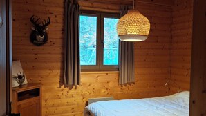 A wooden cabin room with a bed, a small wooden cabinet, a chair, a window with curtains, and a wall-mounted deer head decoration. - Chalet des Etangs in La Bresse for 8 people (La Bresse)