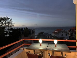 A balcony with two tables and chairs, overlooking a coastal view with buildings and a distant shoreline.
