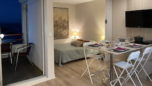 Et moderne hotelværelse med balkon, spiseplads med et bord dækket til to og et stort fladskærms-tv på væggen.