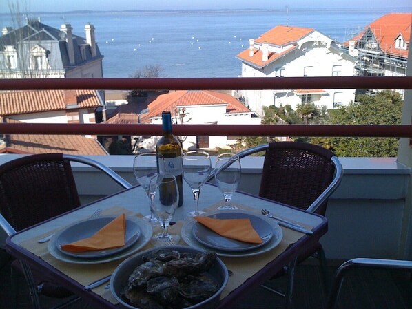 A table set for two with a bottle of wine, glasses, plates, and a bowl of oysters. The table is on a balcony with a view of the sea and buildings in the background.