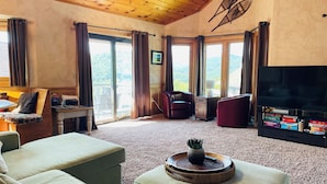 A cozy living room with a flat-screen TV, a coffee table with a plant, and a view of the outdoors through large windows.