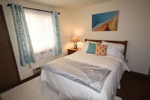 A neatly made bed with a white comforter and pillows, a bedside lamp, and a painting of a beach scene on the wall.
