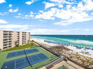 A beachfront tennis court with a view of the ocean and beach umbrellas.