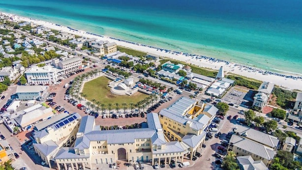 Vista aérea de un pueblo costero con una plaza central, frente al mar y varios edificios, incluyendo un hotel destacado con una estructura en forma de cúpula.