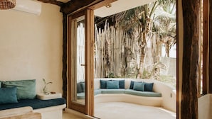 A modern living room with a curved sofa, a wooden bench, and a large glass door leading to a balcony with a tropical view. - Casa Bonita ~ Mahayana, Eclectic Treehouse in the Heart of Tulum Beach (Tulum)