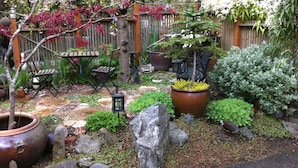 Um jardim com cerca de madeira, plantas em vasos, um banco de pedra e uma pequena mesa com cadeiras.