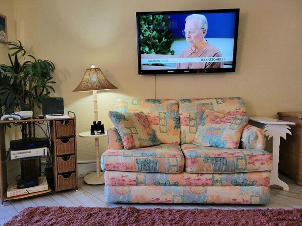 Una sala de estar con un sofá, televisión montada en la pared, una lámpara y una planta.