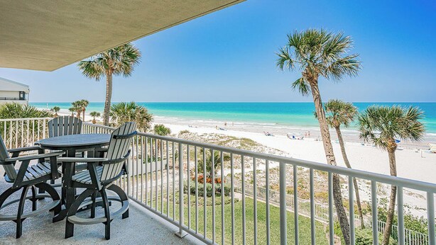 Ein Balkon mit Tisch und Stühlen mit Blick auf einen Strand, an dem Palmen stehen, und einem strahlend blauen Himmel.