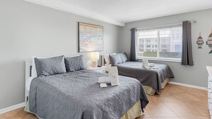 Ein Hotelzimmer mit zwei Betten, einem großen Fenster mit Vorhängen, einem Bild an der Wand und Blick auf Gebäude.