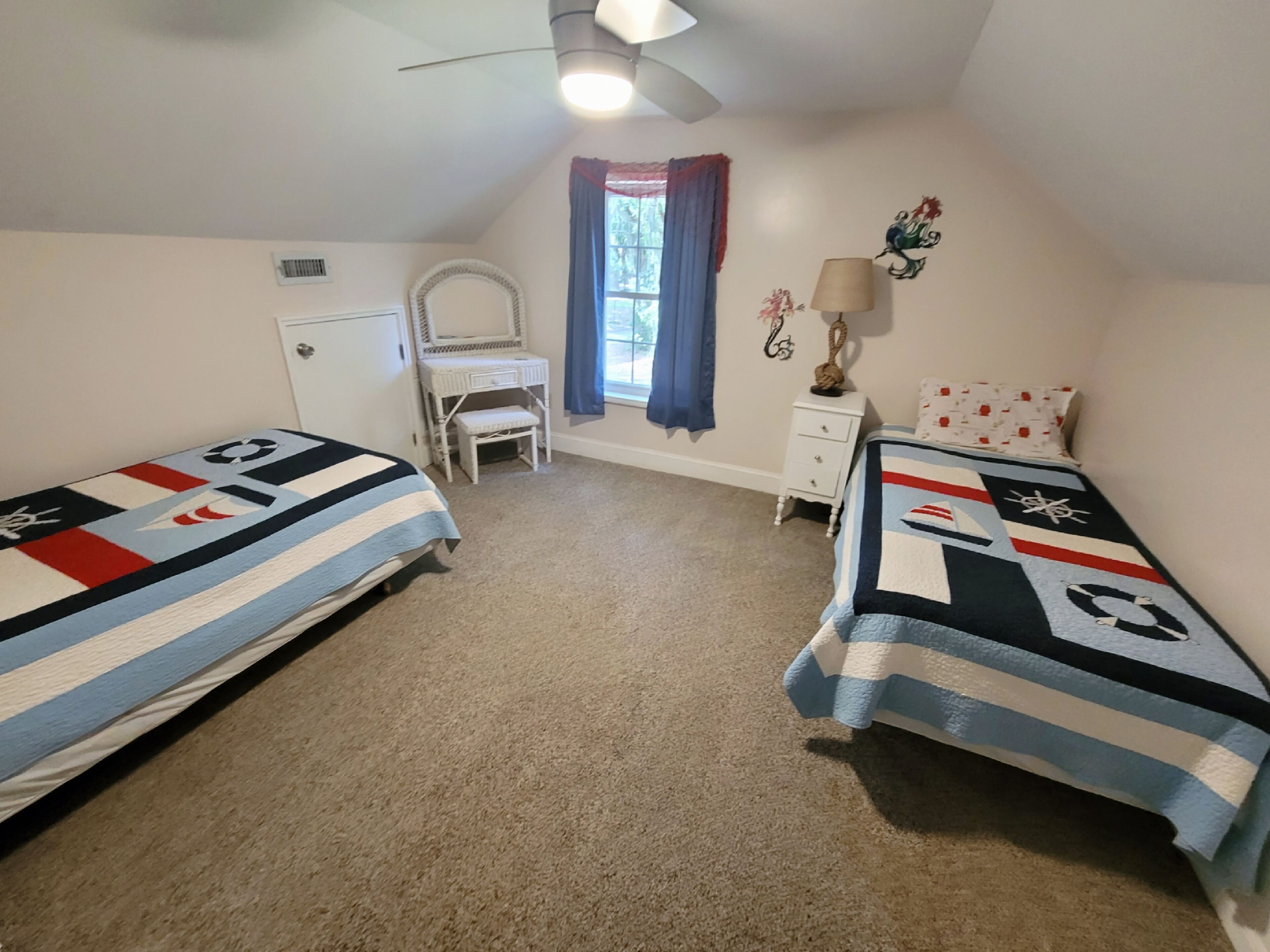 A bedroom with two single beds, a desk, a chair, a small dresser, a mirror, a ceiling fan, and a window with curtains.
