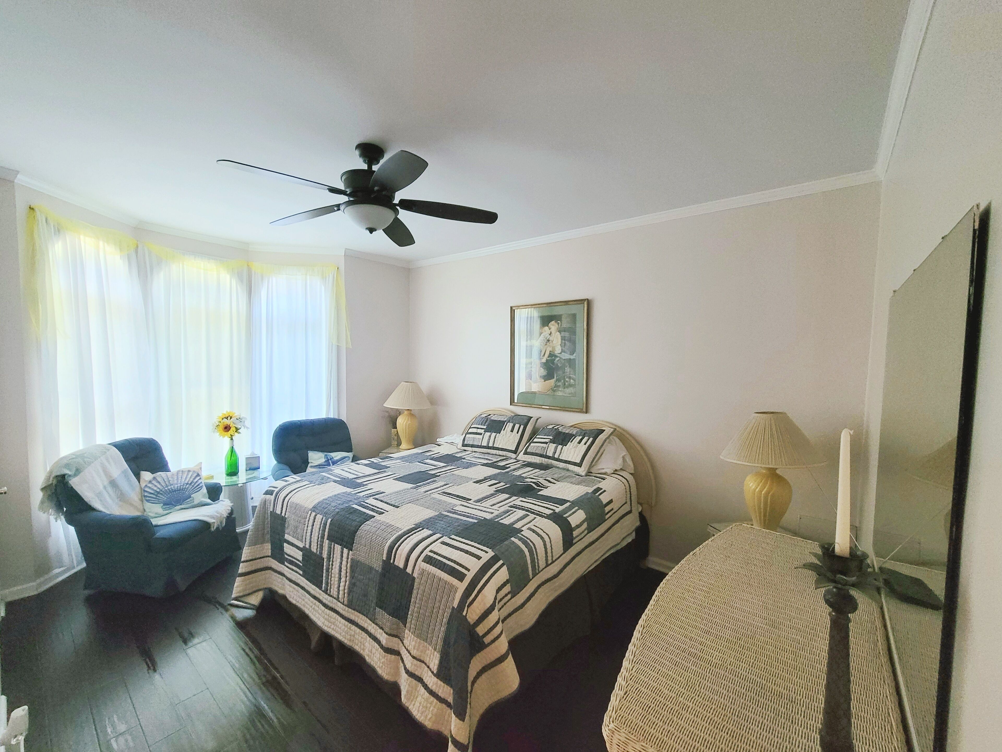 Un dormitorio con cama, una silla, un escritorio, ventilador de techo, ventana con cortinas, lámpara y un cuadro en la pared.