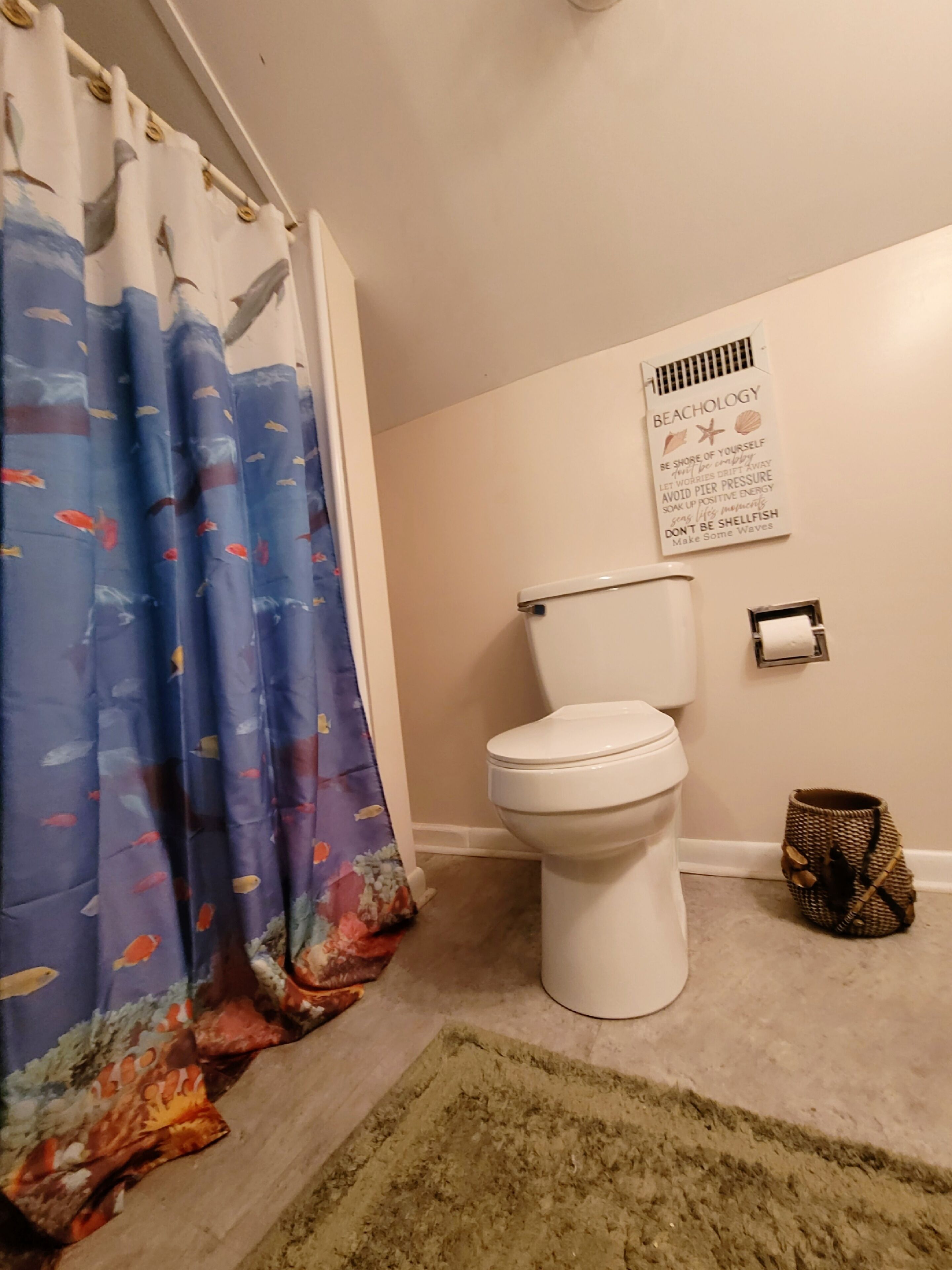 Une salle de bain avec des toilettes, un rideau de douche orné de motifs de poissons, un tapis et un panneau mural avec un texte sur le thème de la plage.