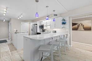 A modern kitchen with white cabinetry, a central island with bar stools, pendant lighting, and a view of a dock through an open wall. - 317 - Fantastic 3 BR Oceanfront Corner (New Smyrna Beach)