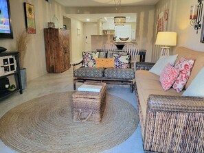 A living room with a sofa, a coffee table, and a TV. There is a kitchen in the background. - 101 - Caribbean décor and a few steps to pool (New Smyrna Beach)