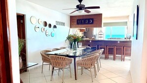 Uma sala de jantar com mesa de vidro, cadeiras de vime, ventilador de teto e cozinha ao fundo.
