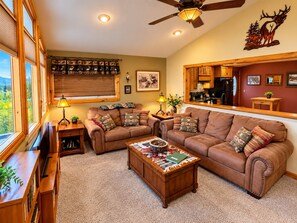 A cozy living room with a fireplace, a ceiling fan, and a large window overlooking a forest. - All-Season Vacation Getaway with Stunning Views! (East Glacier Park)